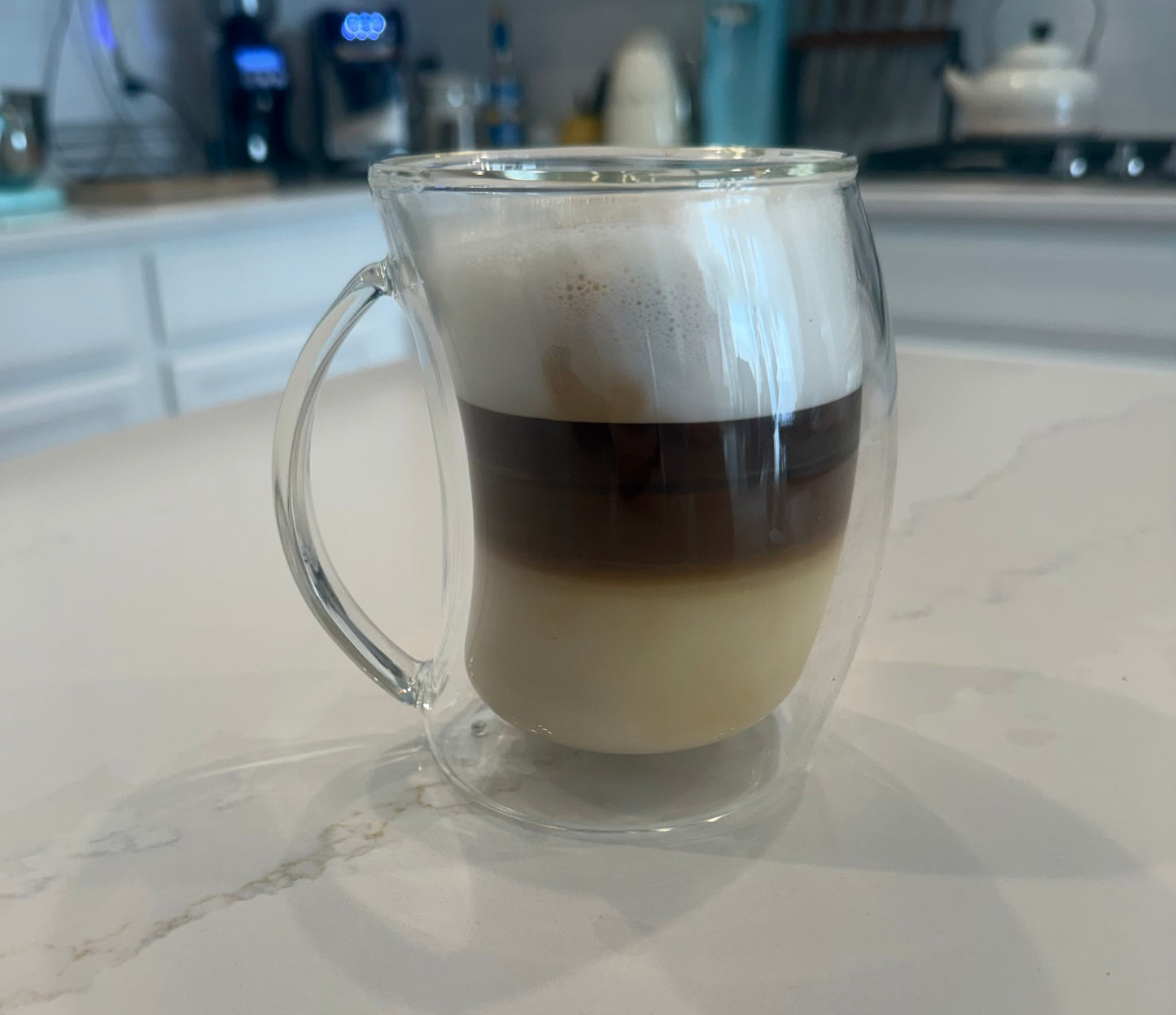 Clear glass mug with layered French Roast brewed coffee and milk on hitches counter
